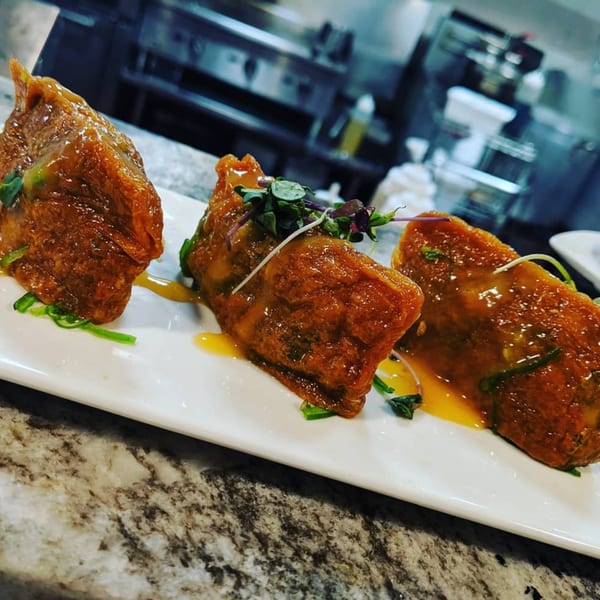 Three glazed Japanese tapas on a white plate with green garnish, in a restaurant kitchen.