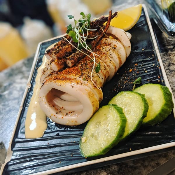 Grilled squid with seasoning, microgreens, lemon, and cucumber slices on a black plate.