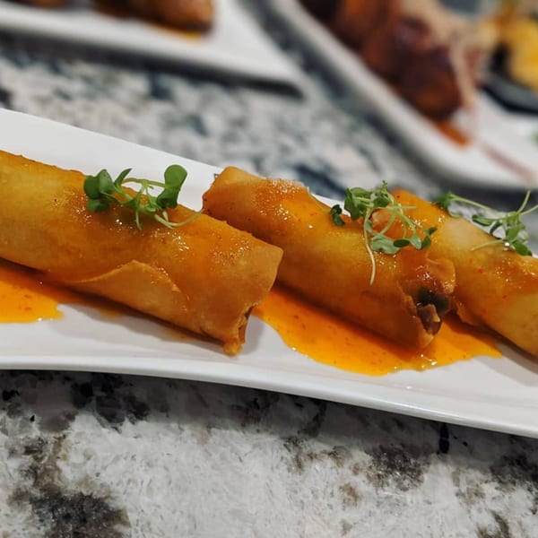 Three golden-brown spring rolls drizzled with orange sauce and garnished with microgreens on a white plate.